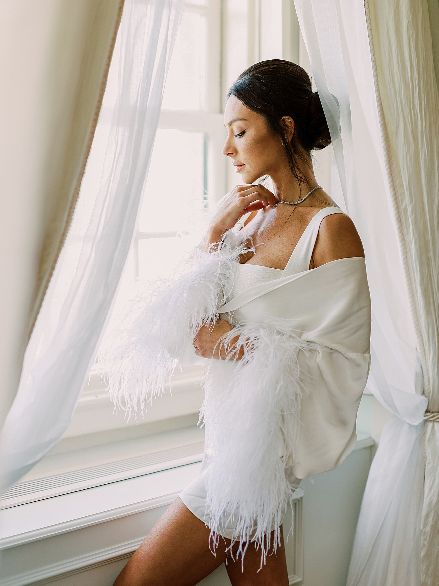 Bride in a robe with fluff sleeves looking out the window | Image by Diane Sotero Photography